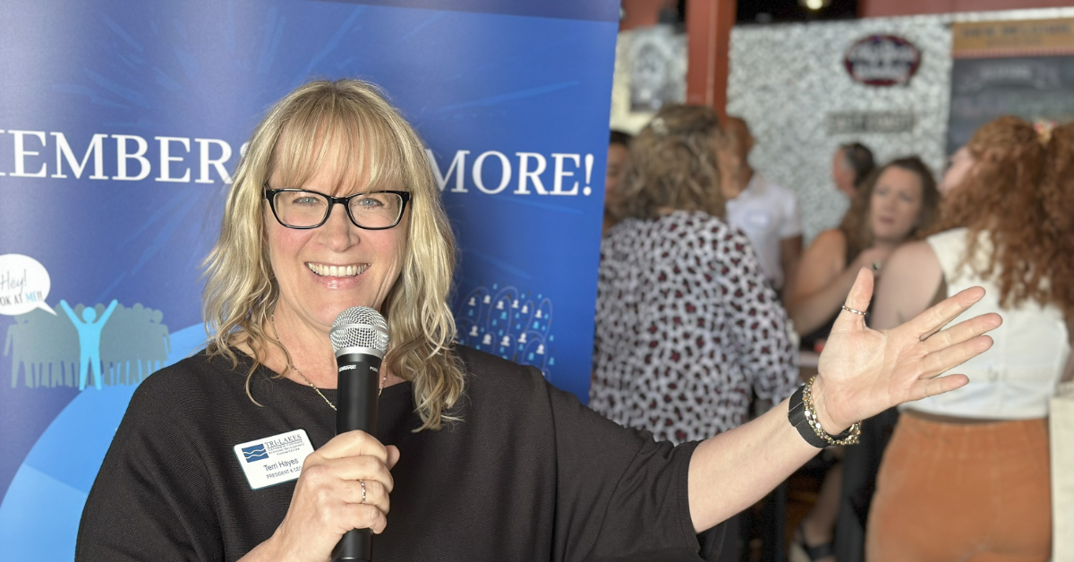 Woman with a microphone speaking at event. Blue banner in background, people mingling.