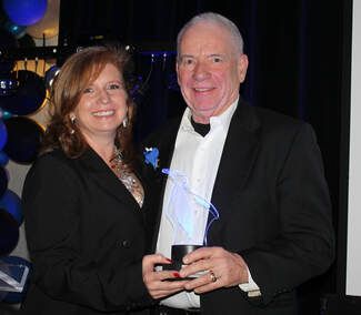 Man and woman smiling, holding an award. Man in a tuxedo, woman in a black blazer, decorated background.