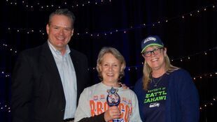 Three people smiling, one holding an award. Two women with the Seattle Seahawks hat and shirt. Man in a suit.
