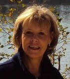 Woman with blonde hair smiling, wearing a dark blue jacket, outdoors near water and trees.
