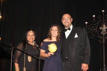 Three people on stage: woman in blue dress holding globe, flanked by a woman in black and a man in a tuxedo.