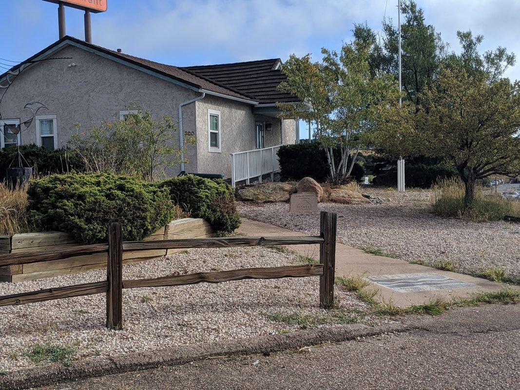 Building with gravel yard, brown wooden fence, shrubs, and a tree, under a cloudy sky.