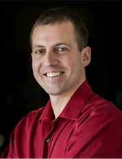 Man in red shirt smiling, upper body shot, against a dark background.