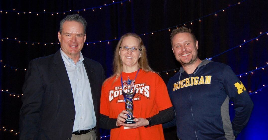 Three people on stage; woman holding trophy, flanked by two men. Blue and orange dominate.