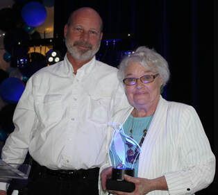 Man and older woman smiling, holding a glowing award. Balloons in background.