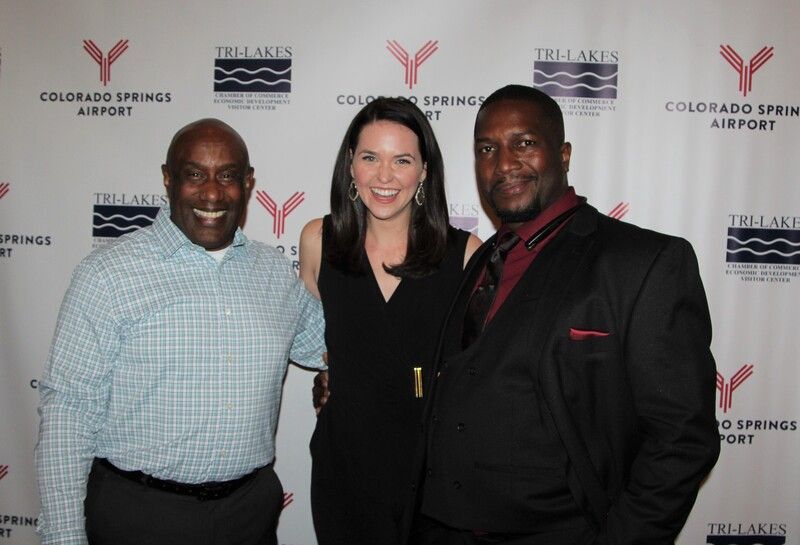 Three people smiling at an event. Two men with a woman, in front of a backdrop with the Colorado Springs Airport logo.