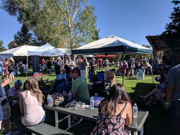 People at an outdoor event under tents, sitting at picnic tables, enjoying drinks, and socializing on a sunny day.