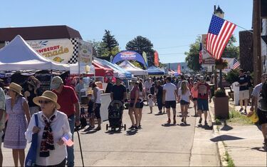 Street fair with crowds, vendors under tents, American flags, and sunny sky.