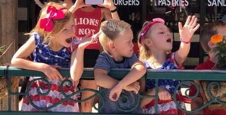Children watching a parade, waving, and wearing patriotic outfits.