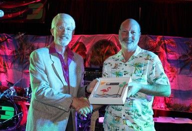 Two men shaking hands, one wearing a suit, the other a tropical shirt, holding a gift box.