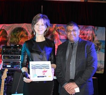 Woman holding an award, smiling, with a man in a suit, indoors, stage backdrop, tropical theme.
