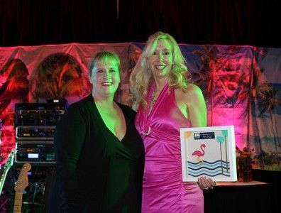 Two women smiling, one in black, the other in a pink dress, holding an award with a flamingo image, on stage.