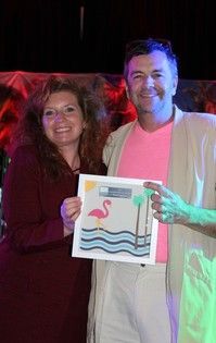 Two people holding a framed artwork with a flamingo. Woman in red smiles, man in pink shirt smiles.