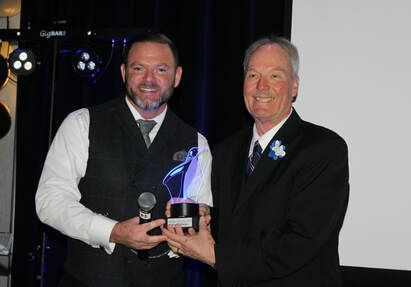 Man in vest holds award, receiving it from another man in a suit at an event.