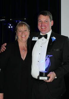 Man in tux holds award next to smiling woman; they pose at a formal event.