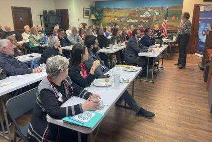 A diverse group of people in a meeting room; a person is lecturing with an American flag in the background.