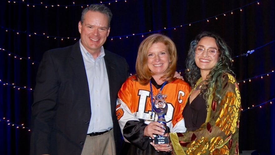 Three people at an event: a man in a suit, woman in a jersey holding an award, and woman in a patterned shawl.