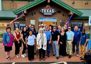 Group of people pose in front of a Texas Roadhouse restaurant; sunny day.