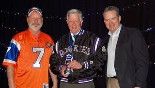 Three men: one in Broncos jersey, one in Rockies jacket holding an award, one in a suit, smiling.