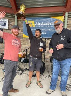 Three men celebrating with a trophy. Outdoors, one raises a trophy, others raise arms. Band backdrop.