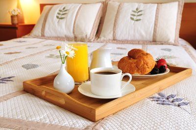 Breakfast tray on a bed with coffee, orange juice, croissant, and flowers.