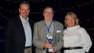 Three people posing; man in center holds award. Blue backdrop.