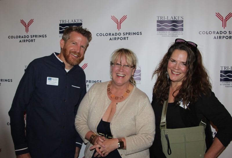 Three people smiling in front of a backdrop with logos for Tri-Lakes and Colorado Springs Airport.