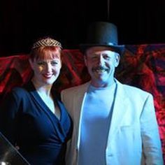 Woman with tiara and man in a top hat, smiling, posing together. Red backdrop.