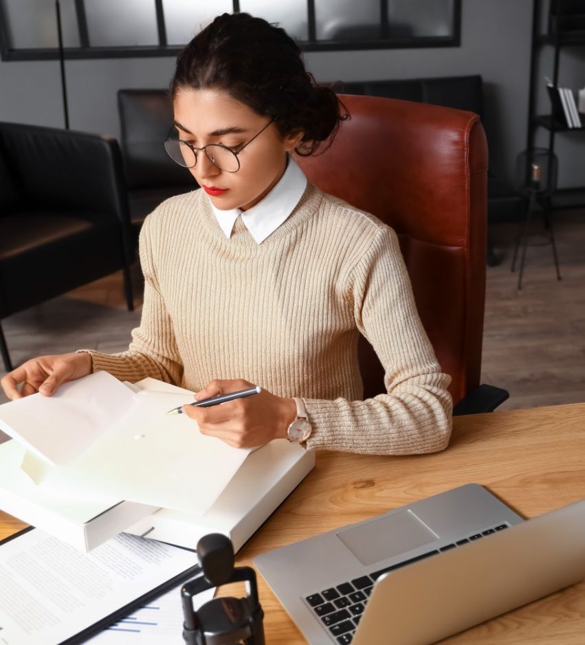 A woman at her desk reviewing paperwork next to an open laptop.