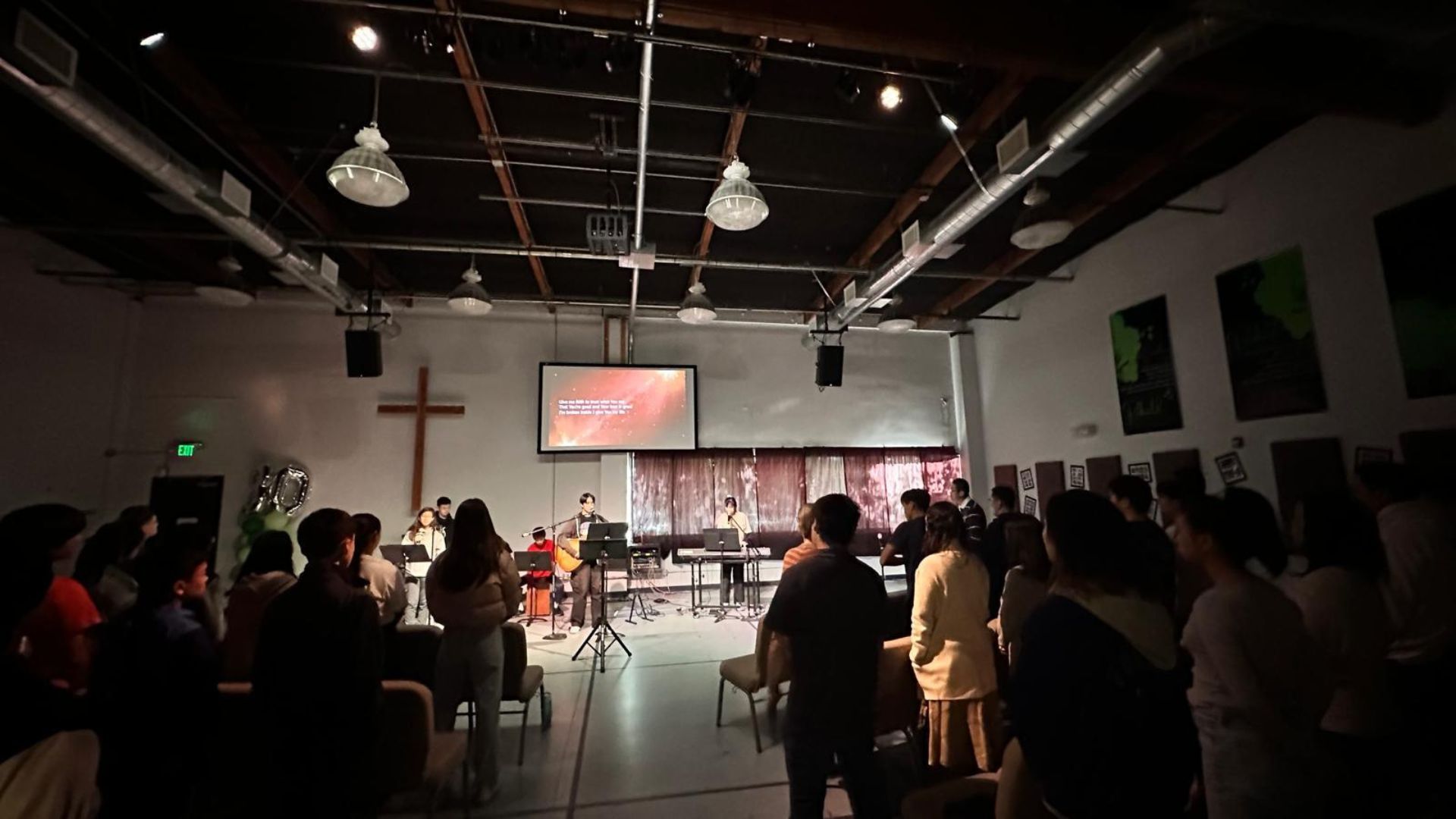 A group stands in a dimly lit, industrial-style room facing a stage with a screen, musical equipment, and a wooden cross.
