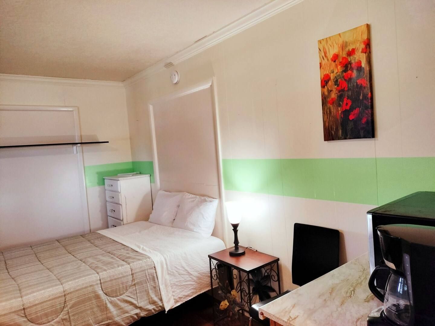 A bedroom with a bed , nightstand , microwave and a painting on the wall.