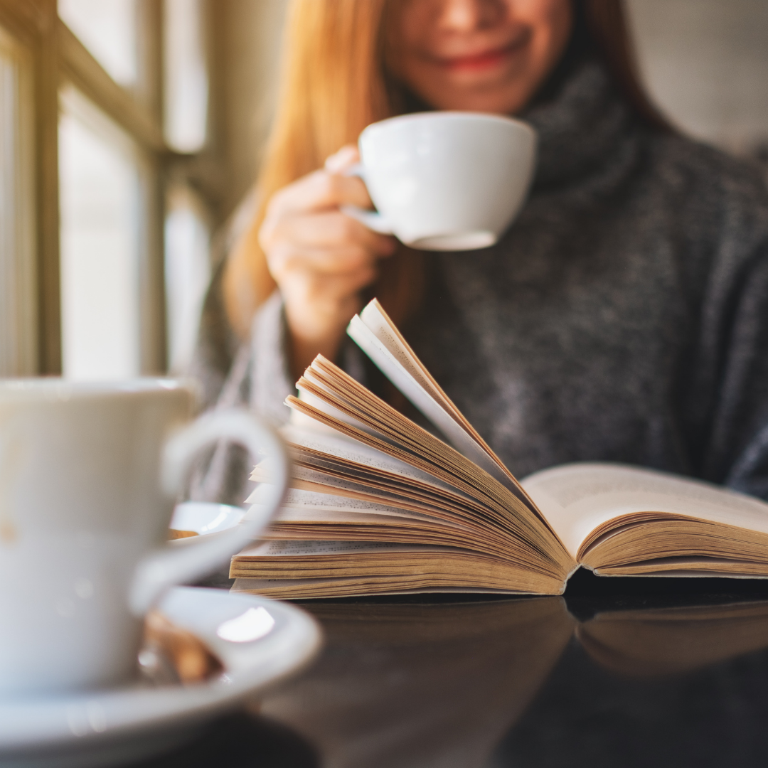 Person holding a cup beside an open book on a table, with a blurred coffee cup in the foreground