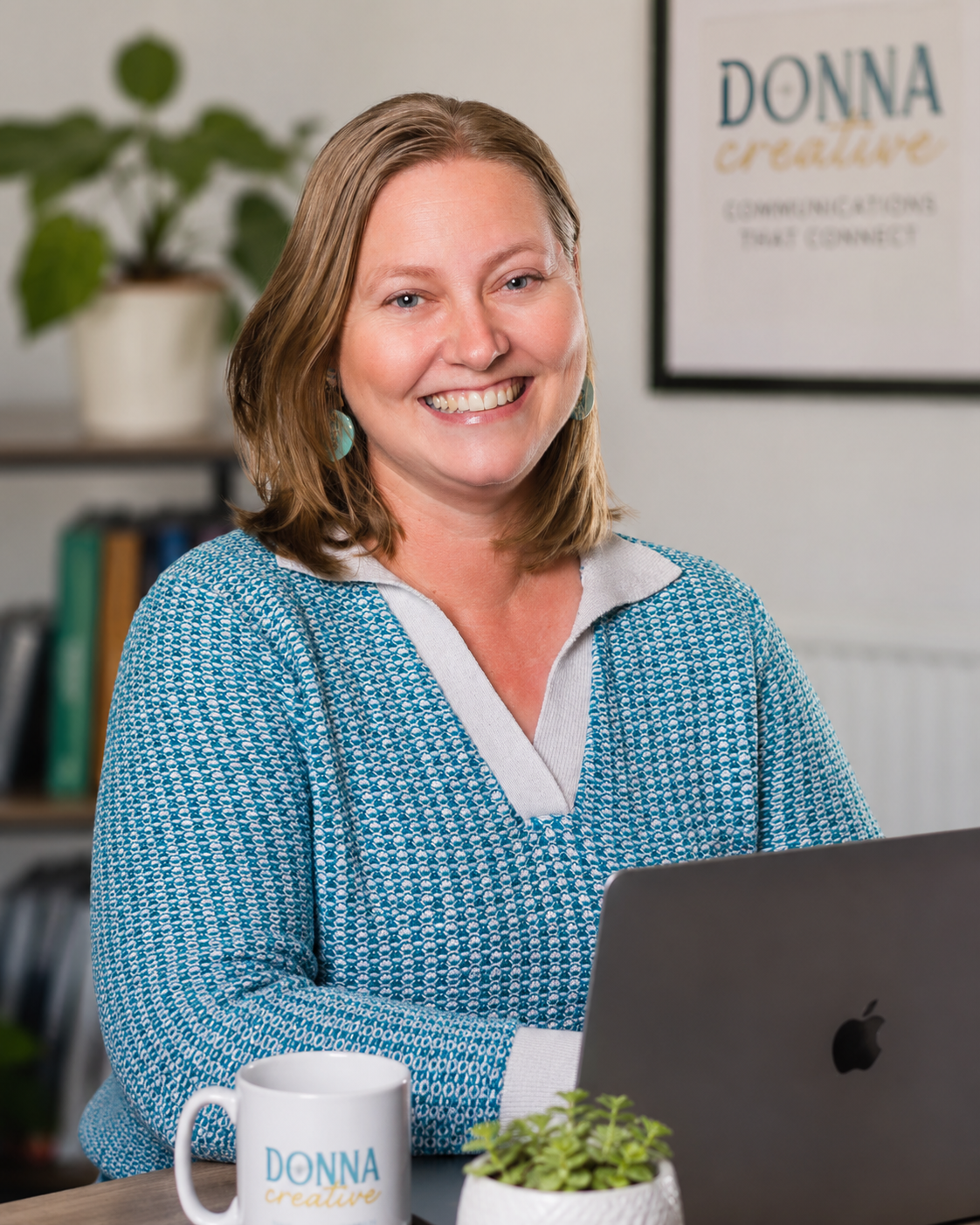Woman smiling at a desk with a laptop, mug, and small plant in an office.