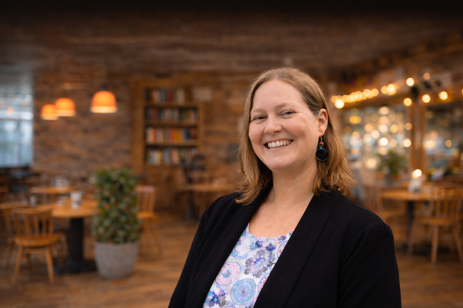 A person smiles in a warm, dimly lit restaurant with wooden tables and a bookshelf in the background.