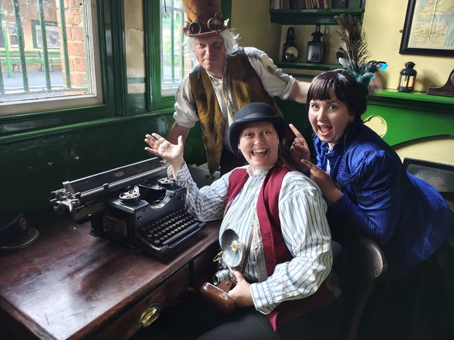Three people dressed in Victorian clothing sat around an old typewriter