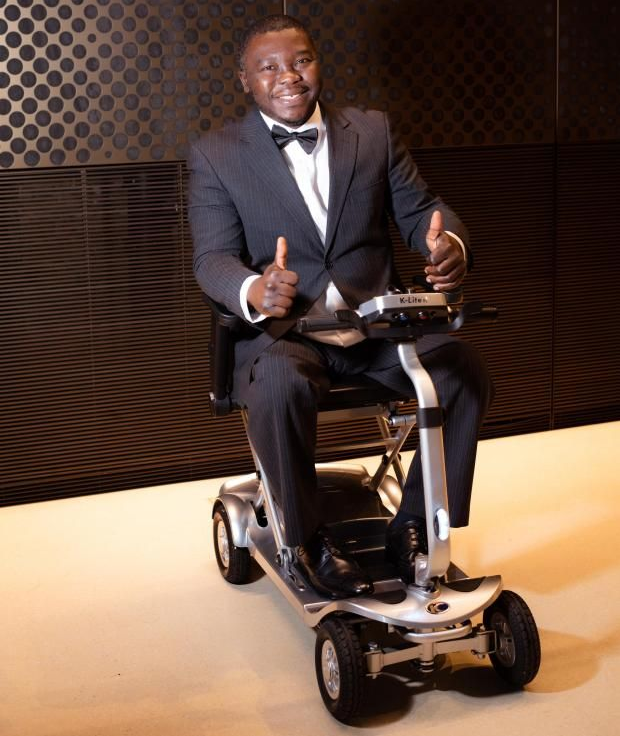 Man in a tuxedo seated on a mobility scooter, smiling in an indoor setting with dark patterned walls