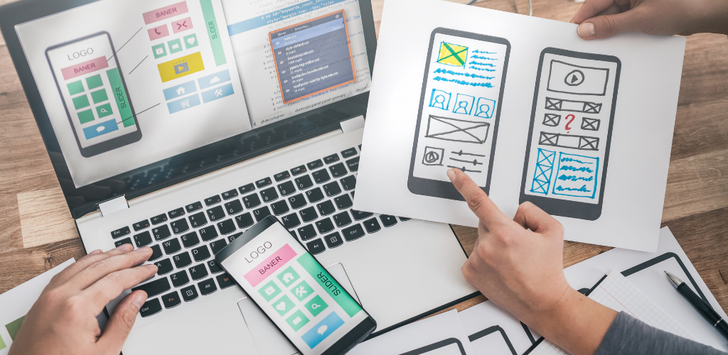 Hands reviewing colorful app wireframes on laptop, tablet, and phone on a desk