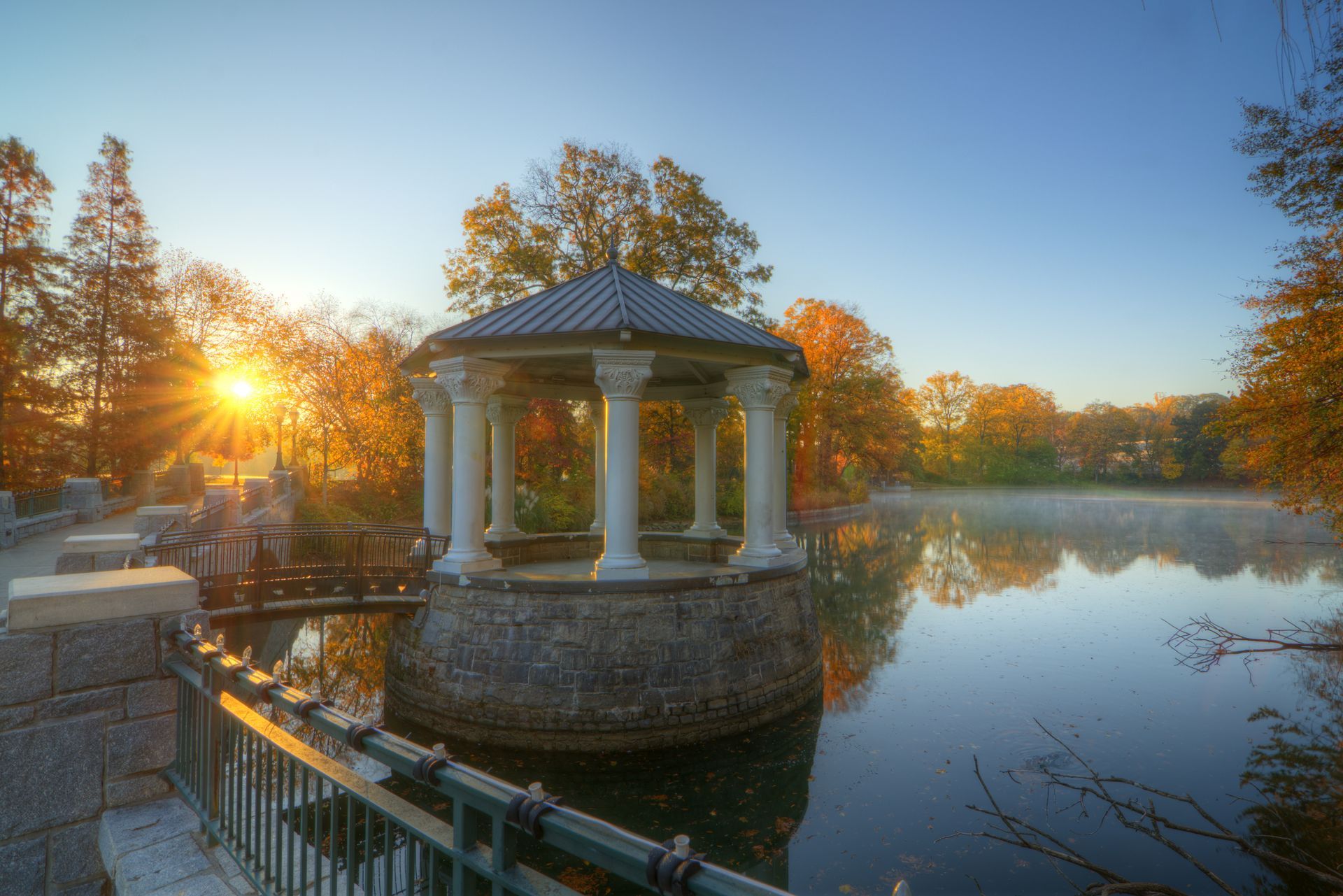Gazebo on a lake at sunrise; surrounded by trees with fall colors.