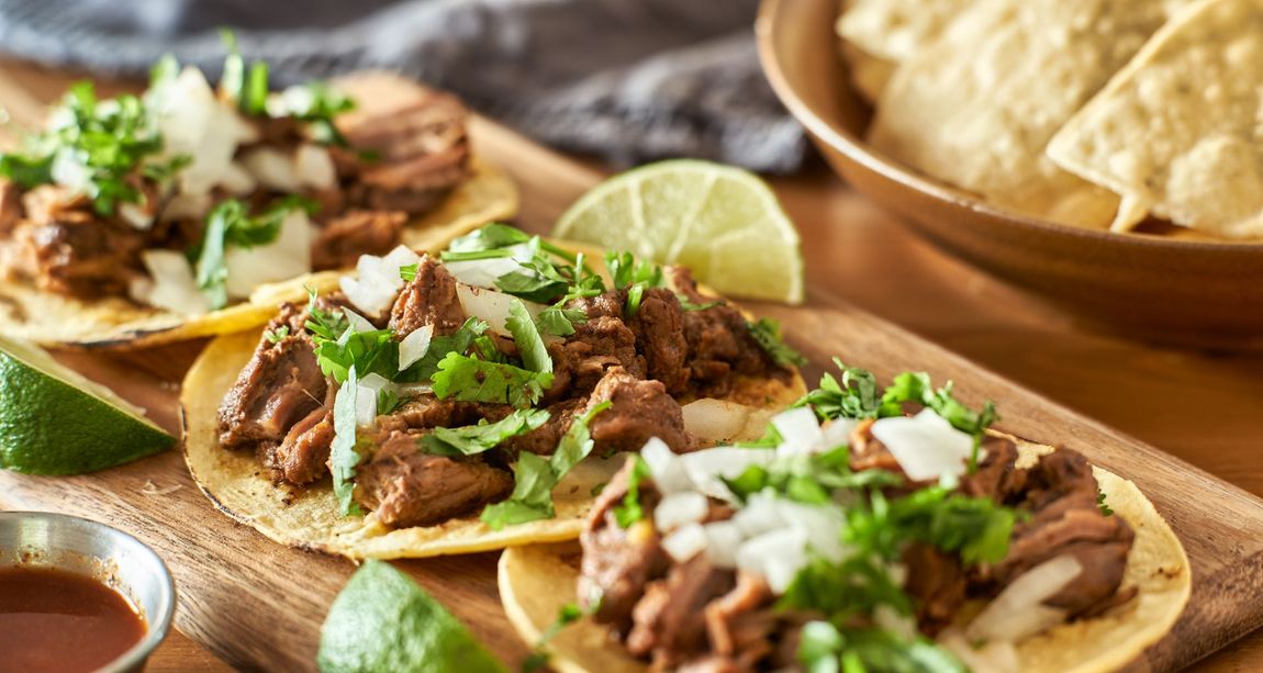 Tacos topped with cilantro, onions, and lime wedges on a wooden board; tortilla chips in a bowl. Tacos topped with cilantro, onions, and lime wedges on a wooden board; tortilla chips in a bowl.