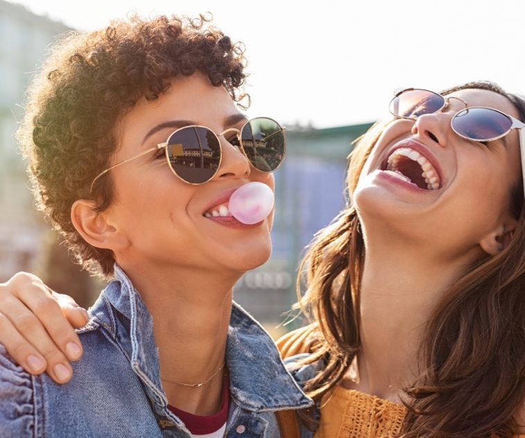 Two women wearing sunglasses are blowing bubble gum and laughing.