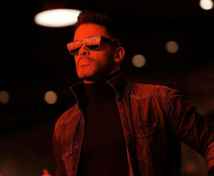 A man wearing sunglasses and a black turtleneck is standing in a dark room.