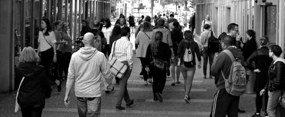 Crowd of people walking down a city street, mostly from the back. Black and white photograph.