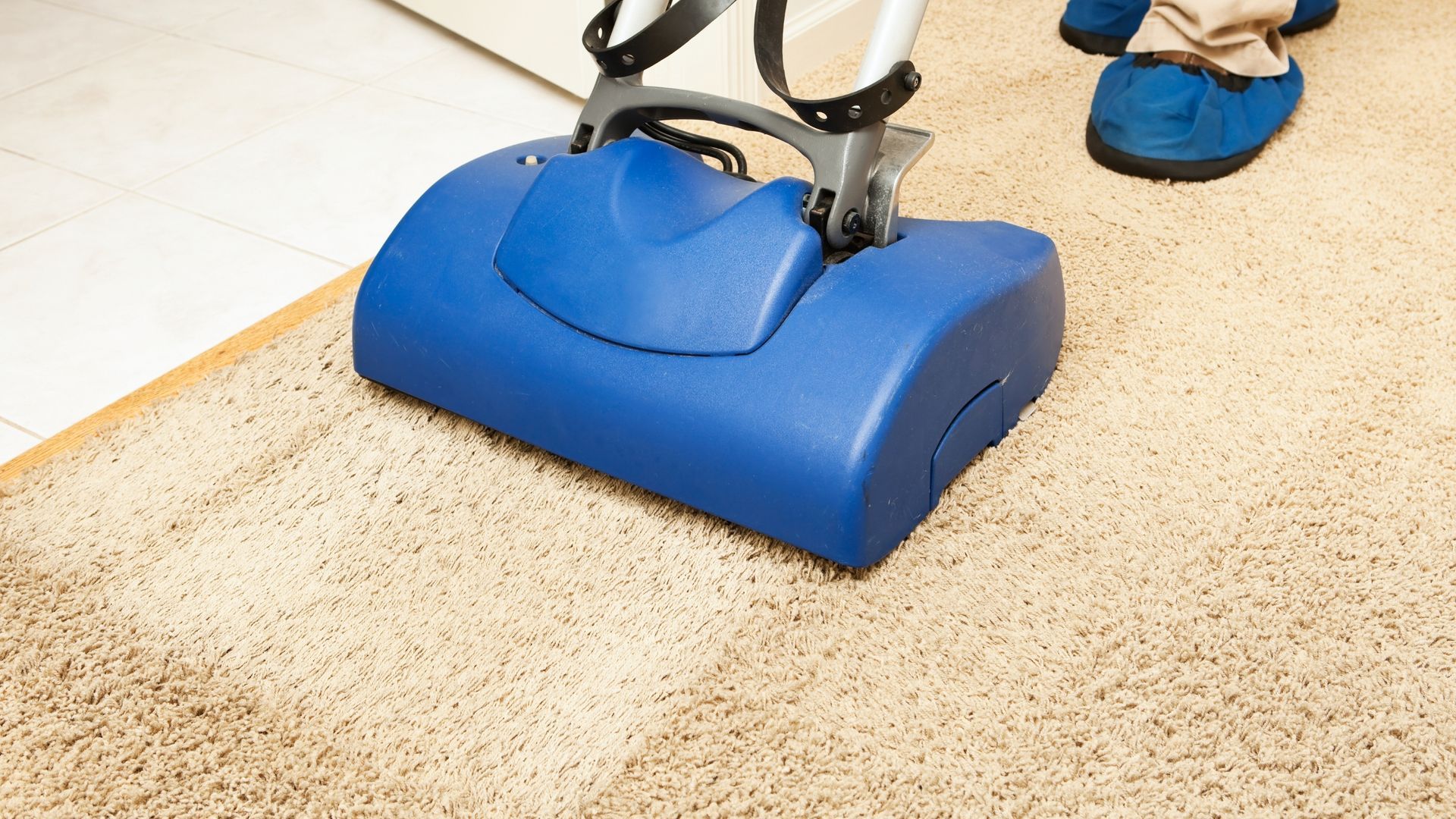 Blue floor scrubber cleaning a beige tile floor, with a person’s feet visible nearby