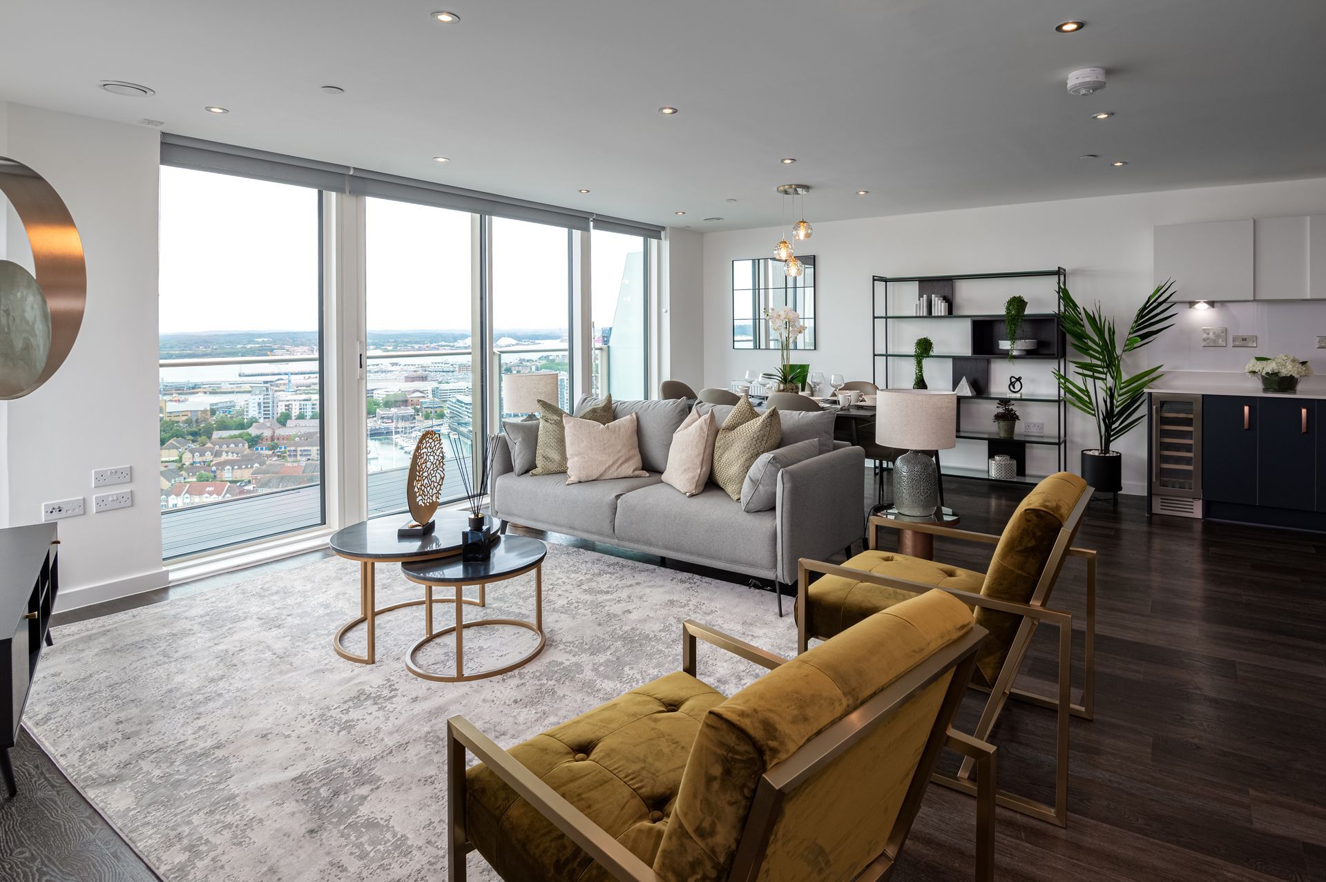 Modern living room with floor-to-ceiling windows, gray sofa, mustard chairs, and dark wood floors overlooking a city view