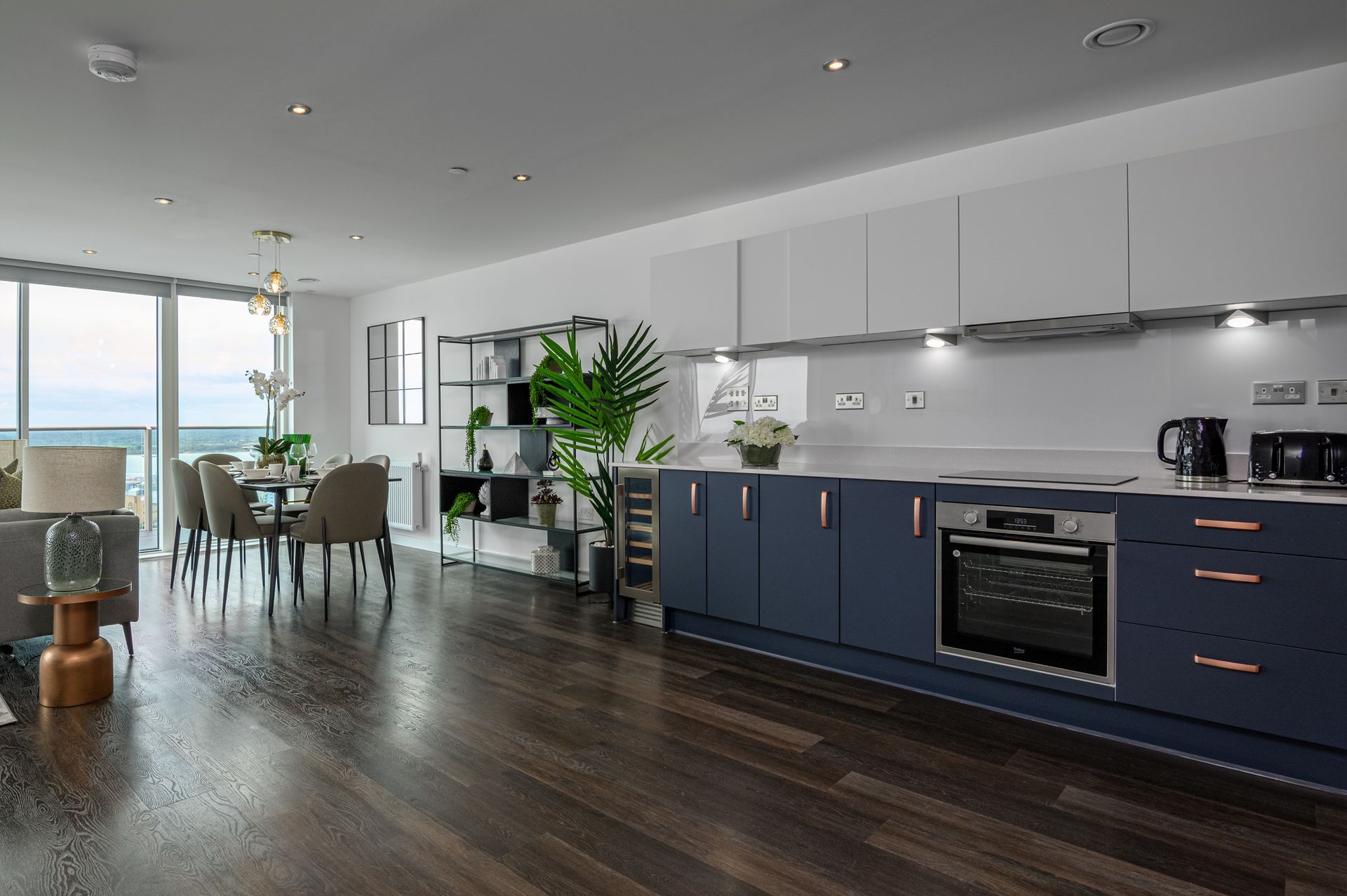 Modern open-plan kitchen and dining area with navy cabinets, dark wood floors, and large windows overlooking water