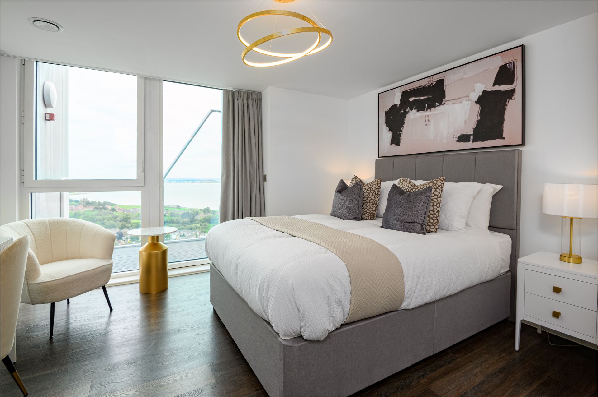 Modern bedroom with a large bed, armchair, bedside table, and floor-to-ceiling window overlooking a city view