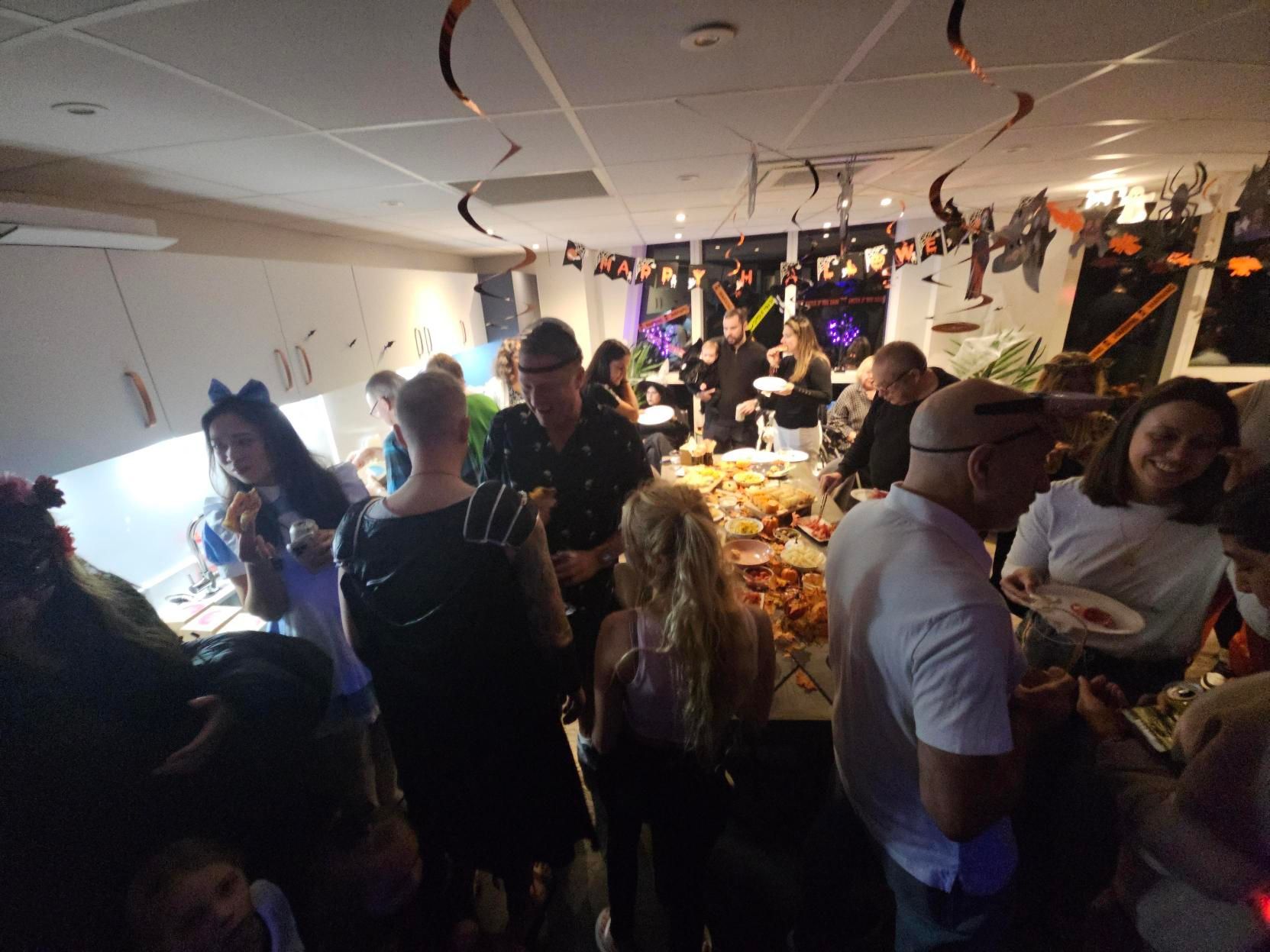 Crowded indoor party with people mingling around a decorated buffet table and hanging streamers