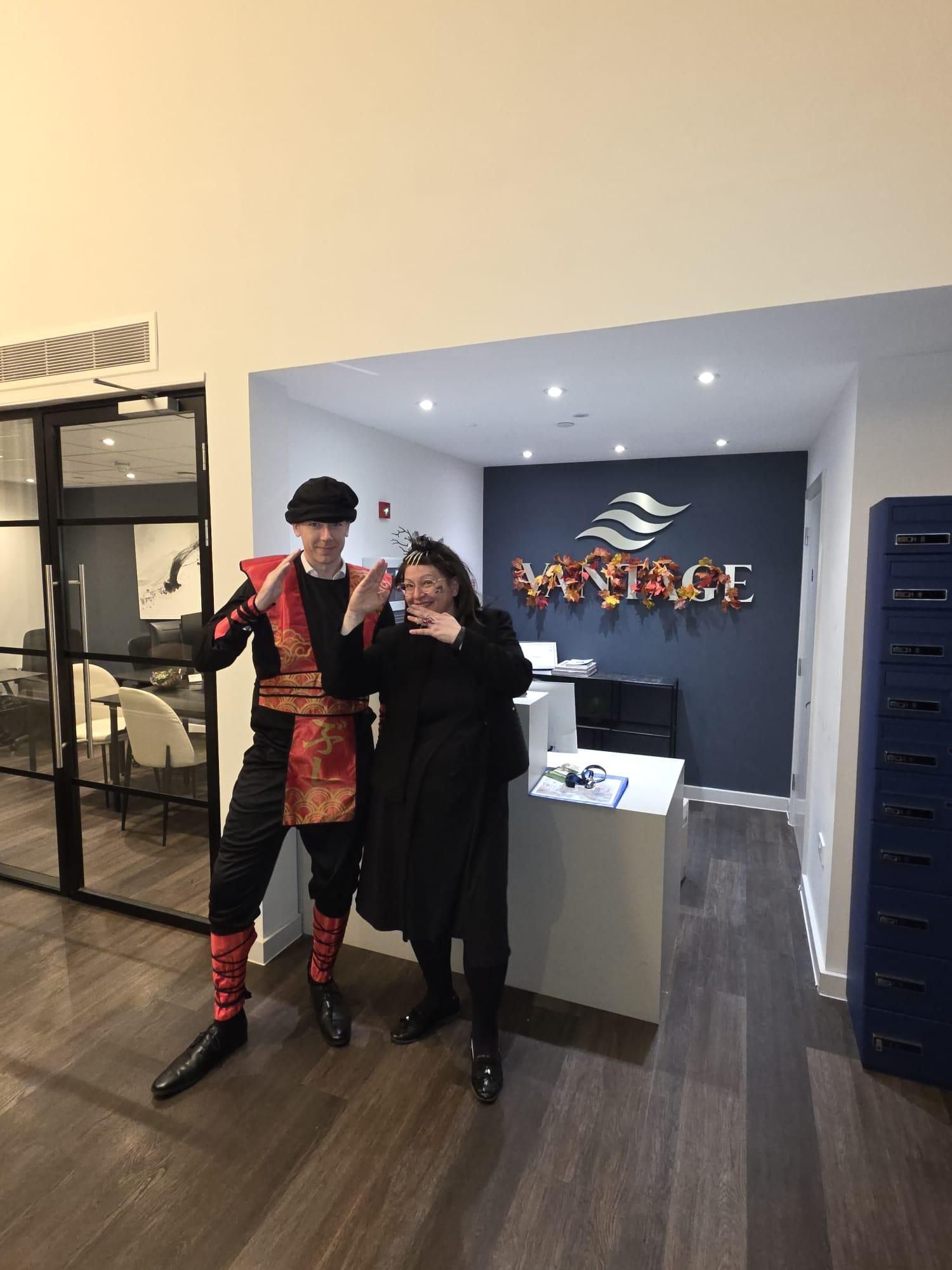 Two people posing in a lobby beside a sign and reception desk, one in a red plaid outfit and black hat, the other in black.