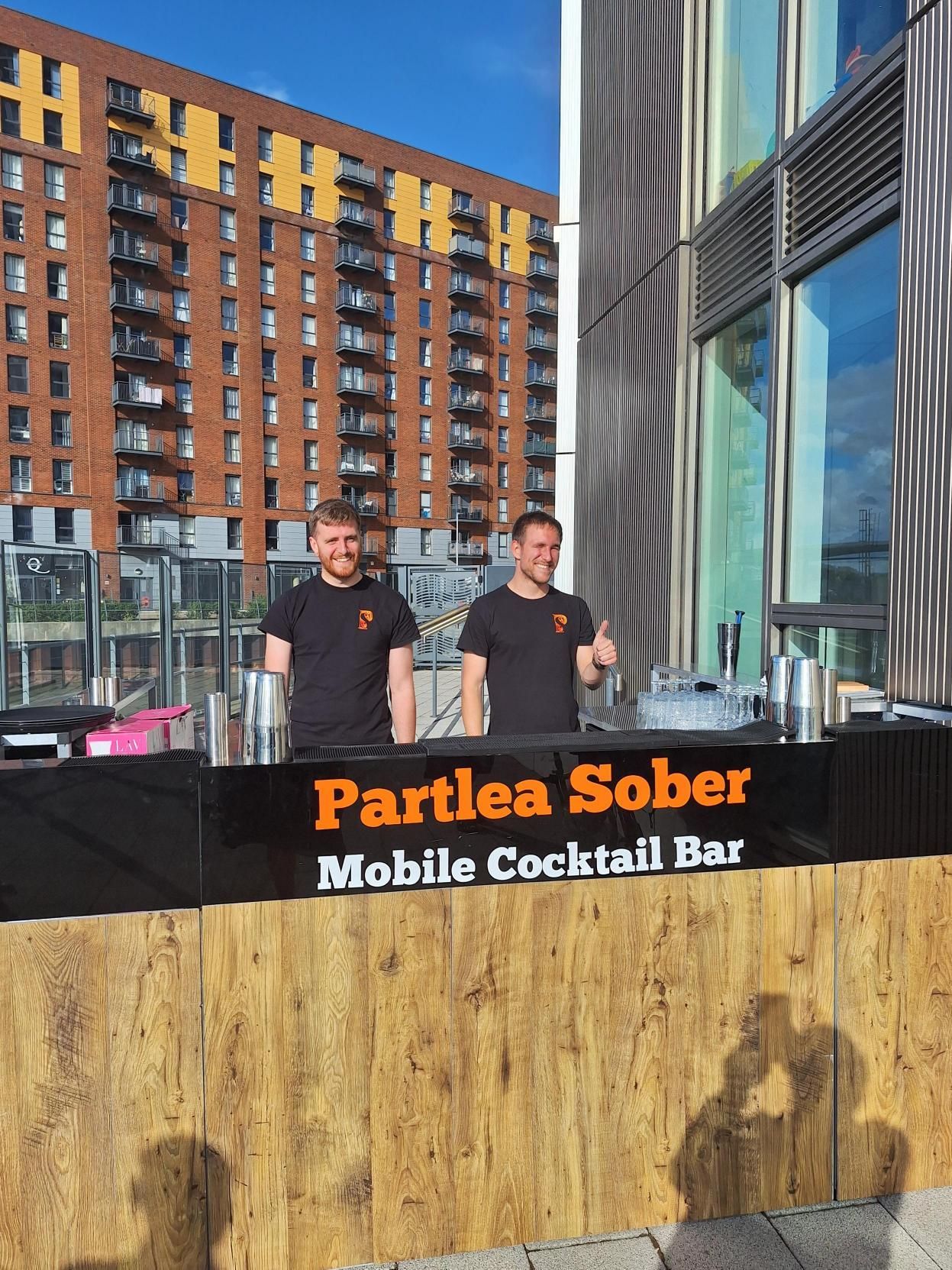 Two men behind a bar labeled “Partlea Sober Mobile Cocktail Bar” in an outdoor urban setting