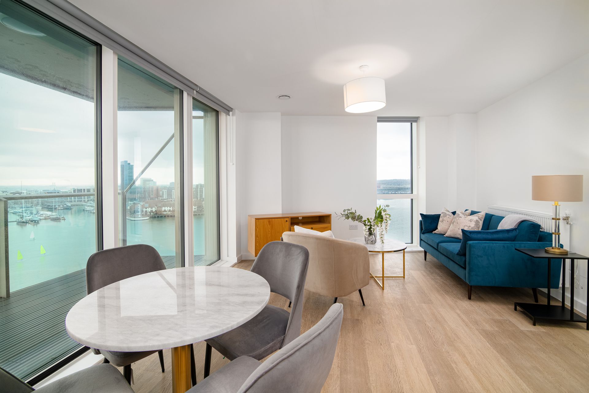 Bright modern living room with large windows, round dining table, blue sofa, and city view